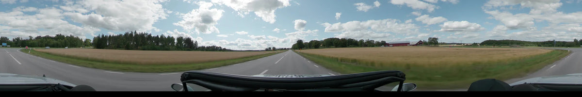 A straight road between Uppsala and Upplands-Bro stretches through vast golden fields and green woodlands under a partly cloudy sky.