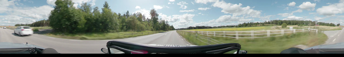 A straight road in Upplands-Bro is bordered by a white wooden fence and open green fields, with dense forests under a partly cloudy sky.