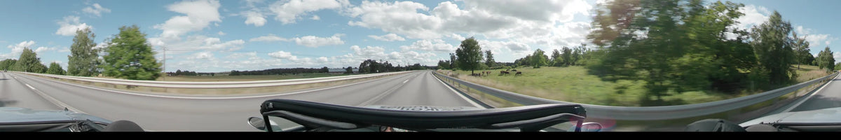 A multi-lane road in Upplands-Bro passes green pastures with grazing cows and scattered trees under a partly cloudy sky.