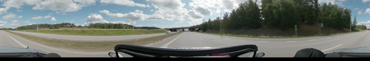 A multi-lane road in Upplands-Bro passes under a concrete bridge, flanked by green grass and dense forests under a partly cloudy summer sky.