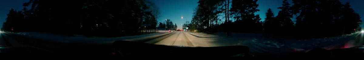 A red car and lit street lamps flank a snowy road in Uppsala, Sweden, on a clear winter afternoon, with dense forests on both sides.