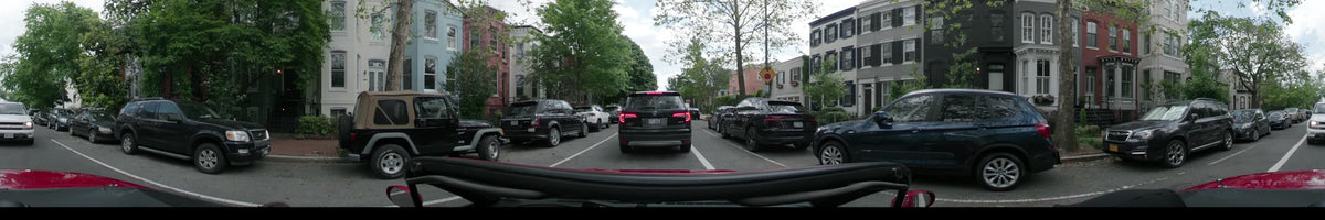 Parked cars and leafy green trees line a residential road in Washington, United States, as a car travels through on a partly cloudy spring afternoon.