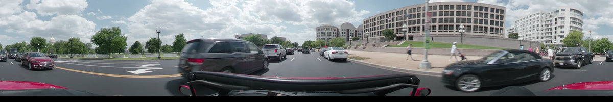 POV shot from a car driving on a city street in Washington during a partly cloudy spring afternoon, surrounded by modern architecture and traffic.