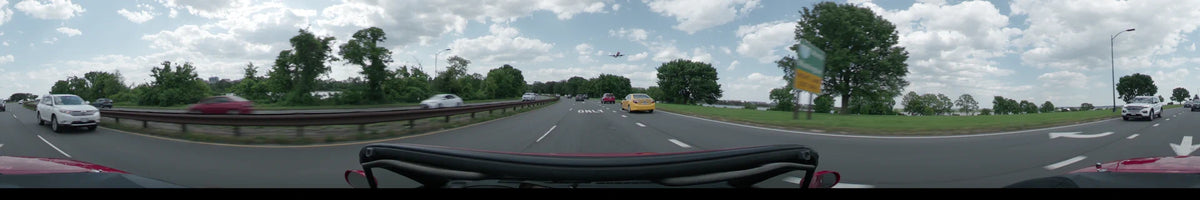 An airplane flies across a partly cloudy sky as a car travels along a multi-lane highway in Washington amidst green spring foliage during the afternoon.