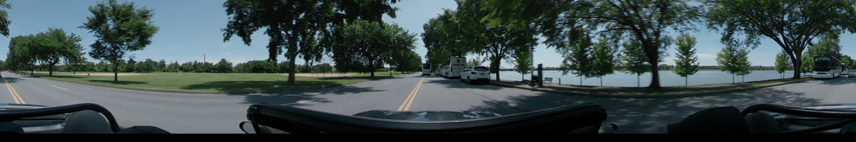 Driving through Washington, United States, on a clear spring day, a car passes white buses and a white car along a road bordered by lush trees and water.