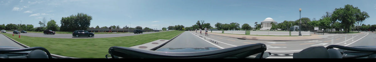 Several cyclists ride ahead of a car on a road in Washington, United States, on a clear spring day, flanked by green grass and trees.