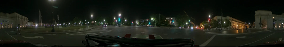 A car moves along an empty road past glowing streetlamps, green traffic signals, and illuminated modern buildings in Washington, United States, on a clear spring night.