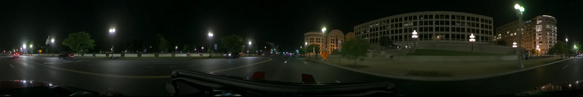 Under glowing streetlamps, a car drives past a yellow road sign and a large, illuminated building with curved facades in Washington, United States, on a clear spring night.
